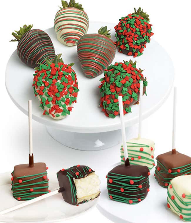 A display of holiday-themed treats featuring chocolate-covered strawberries decorated with red and green sprinkles or drizzles, and chocolate-covered cheesecake bites on sticks with festive red and green decorations.