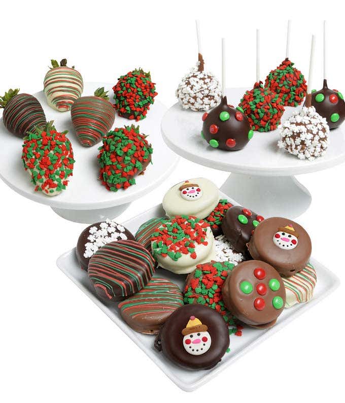 Festive treats including chocolate-covered strawberries with red and green sprinkles, cake pops with holiday decorations, and various decorated chocolate-covered cookies. Arranged on white stands and plates for a holiday presentation.