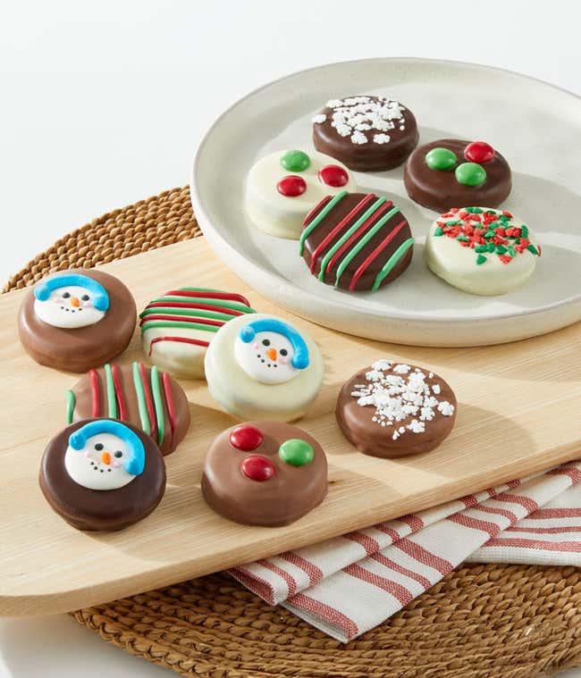 A festive assortment of chocolate-covered cookies decorated with holiday-themed designs, including snowman faces, colorful sprinkles, and red and green candies, arranged on a white plate.