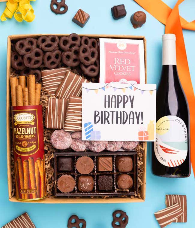 A gift box with chocolate pretzels, chocolate graham crackers, red velvet cookies, hazelnut wafer rolls, an 8 piece box of chocolates, a bottle of red wine and a BIrthday message card.