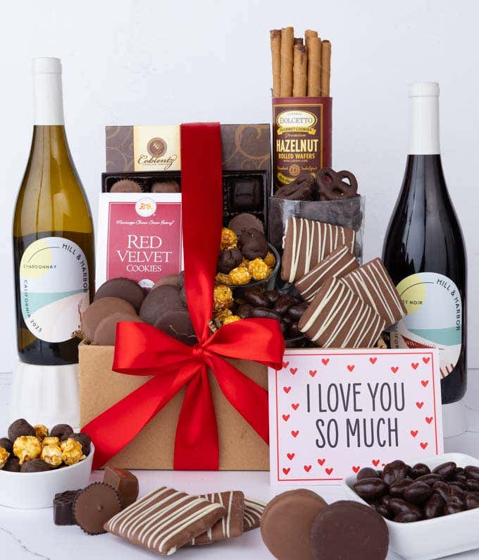 A gift box overflowing with treats including an assortment of chocolate-covered items like oreoes, almonds graham crackers and pretzels along with red velvet cookies, hazelnut wafers, an 8 piece box of chocolates, 2 bottles of wine and an I Love You card.