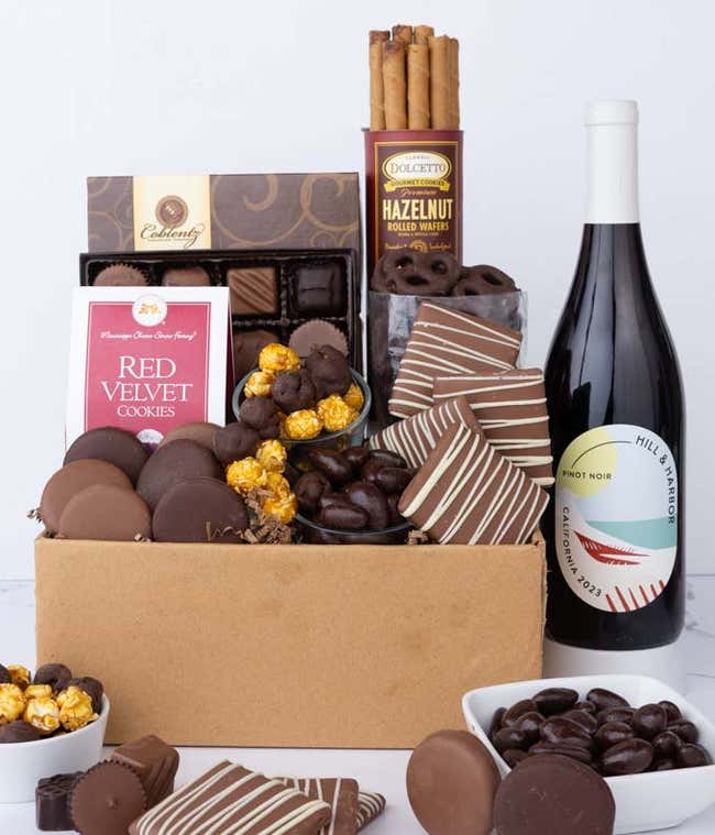 A gift box overflowing with treats including an assortment of chocolate-covered items like oreoes, almonds, graham crackers and pretzels along with red velvet cookies, hazelnut wafers, an 8 piece box of chocolates and a bottle of red wine.