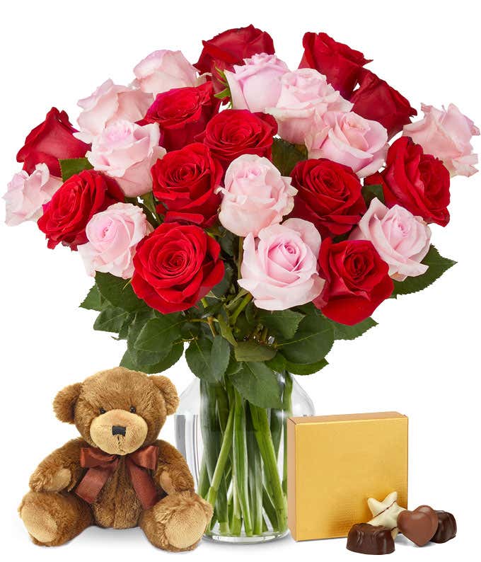 A bouquet of red and pink roses with lush greenery arranged in a clear glass vase. Sitting next to it is a brown teddy bear and a square gold box of chocolates.