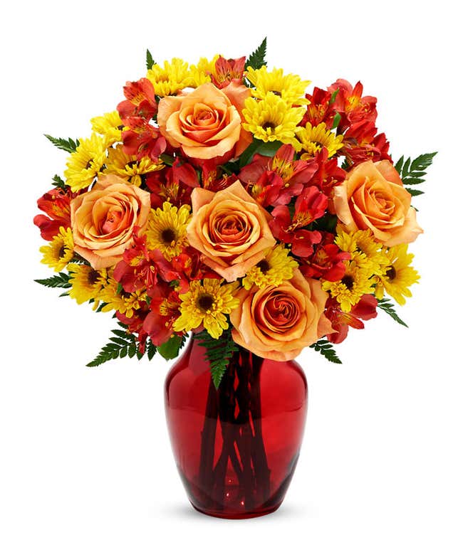 A bright fall bouquet of orange roses, red alstroemeria, and yellow daisies with greens in a glass vase.