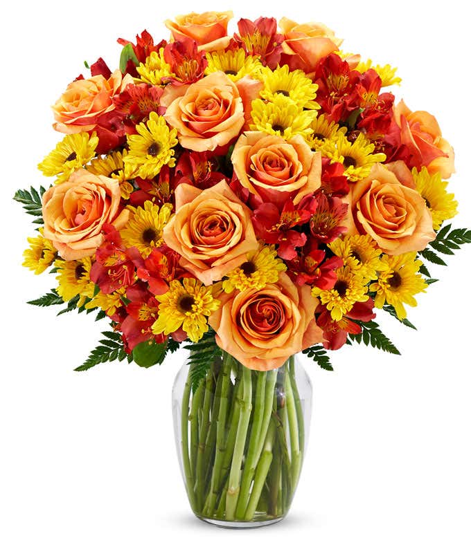 A bright fall bouquet of orange roses, red alstroemeria, and yellow daisies with greens in a glass vase.