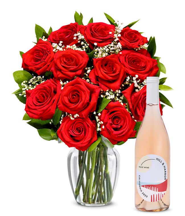 A bouquet of vibrant red roses arranged in a clear glass vase, surrounded by small white flowers and lush green leaves, paired with a bottle of rose wine from California, 2023, featuring a stylish, minimalist label.
