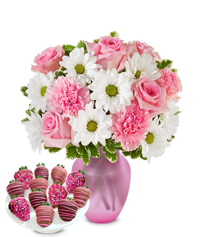 Charming bouquet of pink roses, pink carnations, and white daisies in a clear glass vase, paired with chocolate-covered strawberries decorated with pink drizzle and pink sprinkles.