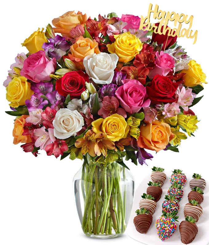 Colorful birthday flower bouquet with mixed roses and alstroemeria in a clear glass vase, topped with a gold “Happy Birthday” pick, paired with an assortment of chocolate-covered strawberries decorated with sprinkles and drizzles.