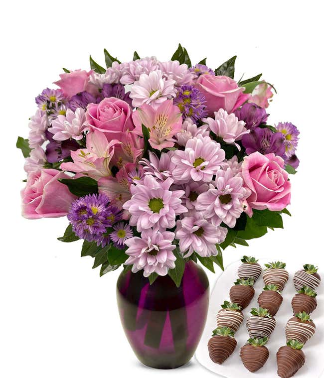 Elegant bouquet of pink roses, lavender cushion poms, purple alstroemeria, pink alstroemeria, and purple asters in a clear glass vase, paired with swizzled chocolate-covered strawberries.