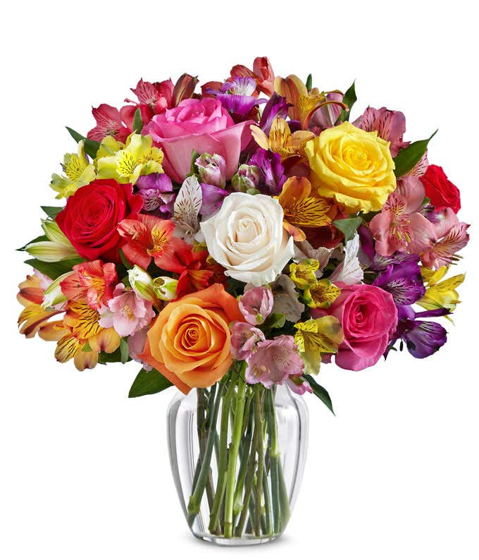 A bouquet of multicolored roses and alstroemeria in shades of red, pink, yellow, orange, and white arranged in a clear glass vase.