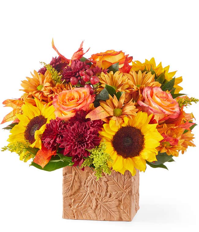 Bright autumn floral arrangement featuring sunflowers, orange roses, red poms, and golden-yellow daisies, arranged in a textured cube vase with a leaf pattern.