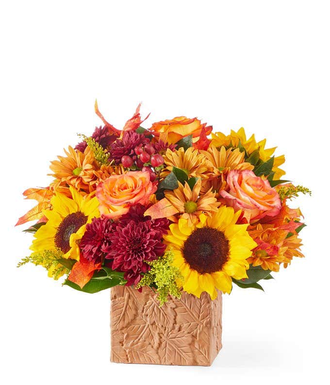 Bright autumn floral arrangement featuring sunflowers, orange roses, red poms, and golden-yellow daisies, arranged in a textured cube vase with a leaf pattern.