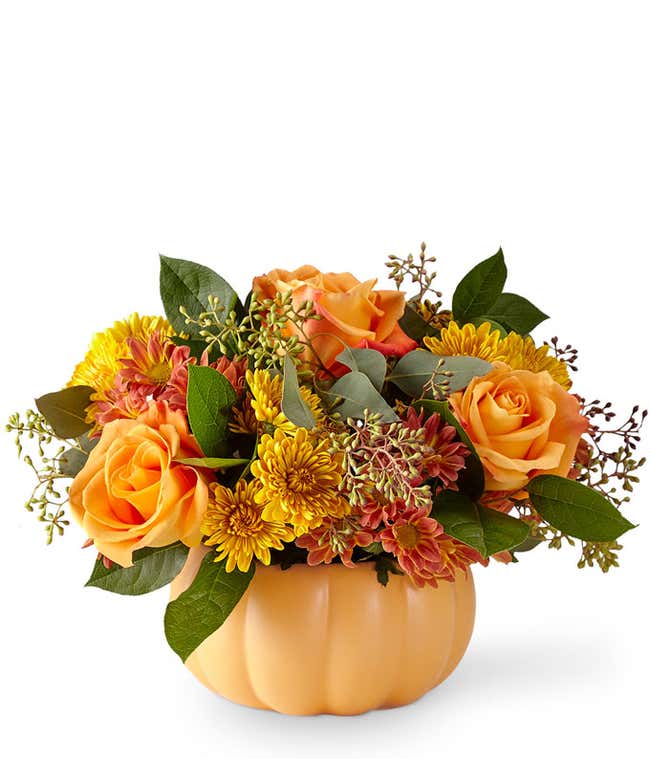  A warm and festive floral arrangement featuring butterscotch cushion poms, bronze daisies, orange roses, and seasonal greens, all beautifully presented in a ceramic pumpkin.
