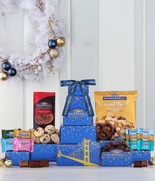 Ghirardelli-themed gift tower with blue boxes featuring golden bridge designs. Contains an assortment of Ghirardelli chocolates, including squares, minis, hot cocoa mix, caramel popcorn, cookies, and chocolate-covered almonds, tied with a Ghirardelli ribb