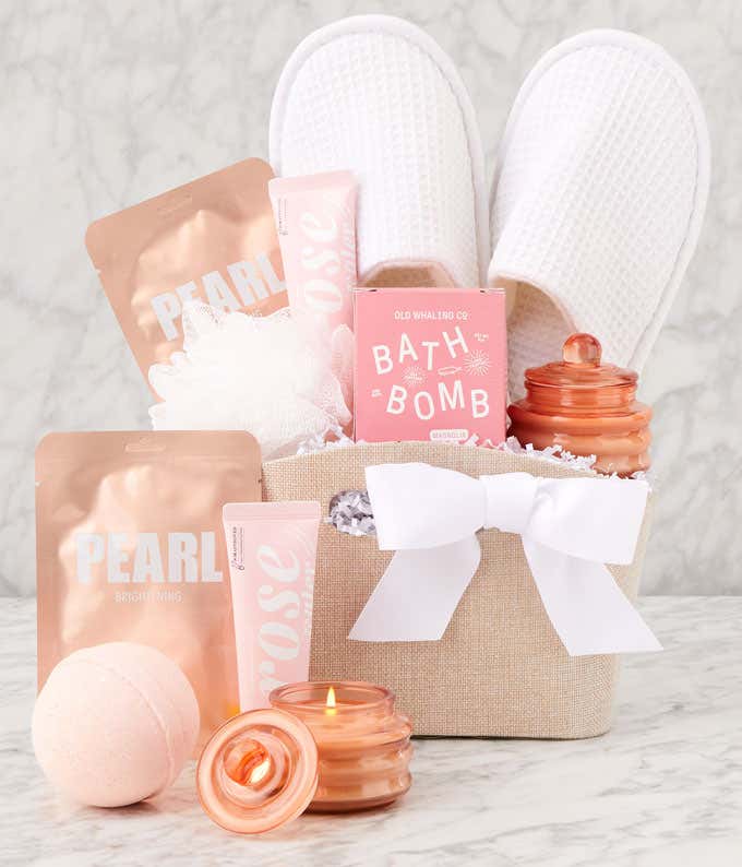 Spa gift set in a beige fabric basket with a white bow, featuring white slippers, bath bombs, pearl face masks, rose-scented lotion, a white loofah, a pink candle, and a pink glass jar, displayed on a marble surface.