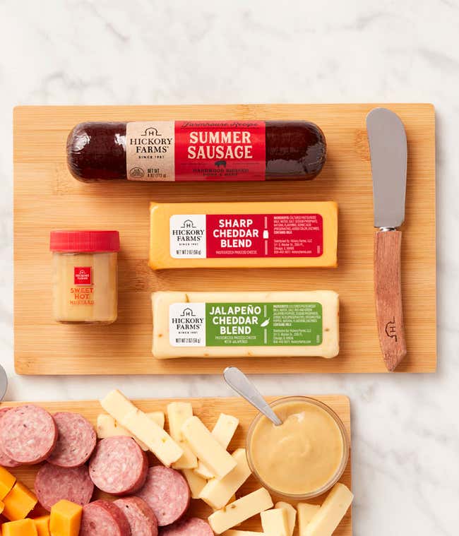Two wooden boards with sliced summer sausage, cubes of sharp and jalape&ntilde;o cheddar cheese, sweet hot mustard, and cheese spreaders, with the full sausage, cheese blocks, and mustard jar displayed above.