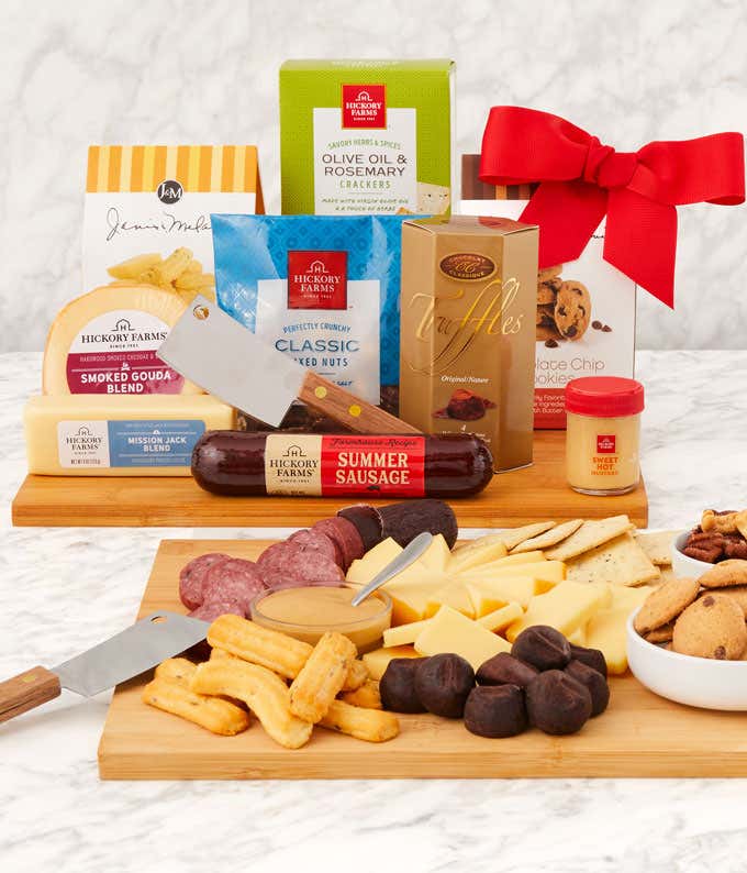 Gourmet gift set with summer sausage, smoked Gouda and cheddar cheese blends, sweet hot mustard, crackers, mixed nuts, chocolate truffles, cookies, and a meat cleaver, displayed on a wooden cutting board with a red bow on one of the boxes.