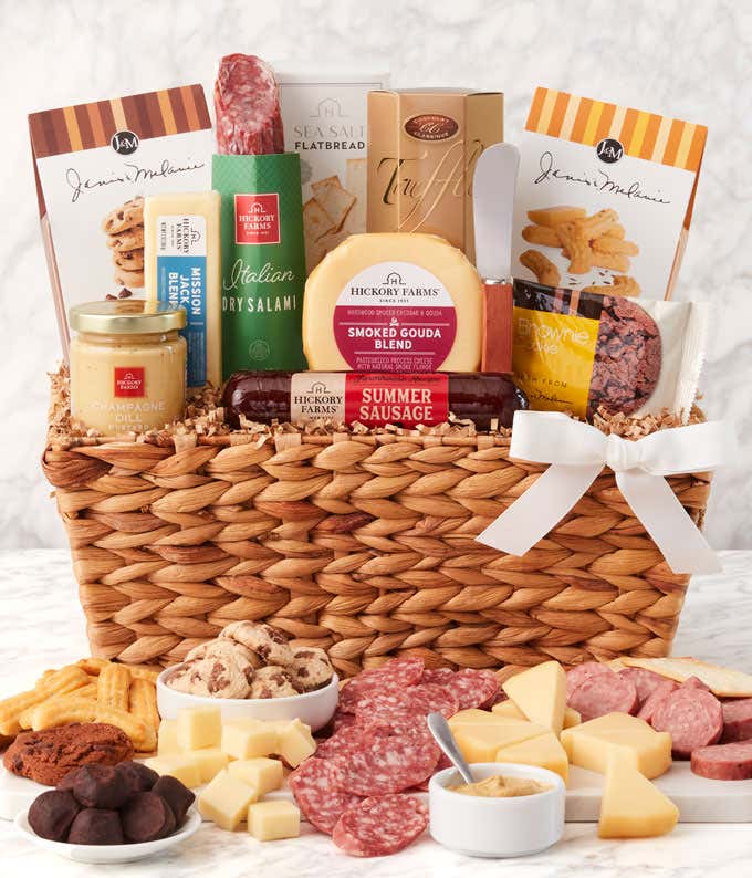 Woven gift basket with salami, cheese, mustard, crackers, cookies, and chocolates. In front, a board displays sliced meats, cheese cubes, and mustard for dipping, with cookies and chocolate truffles arranged alongside. Decorated with a white bow.