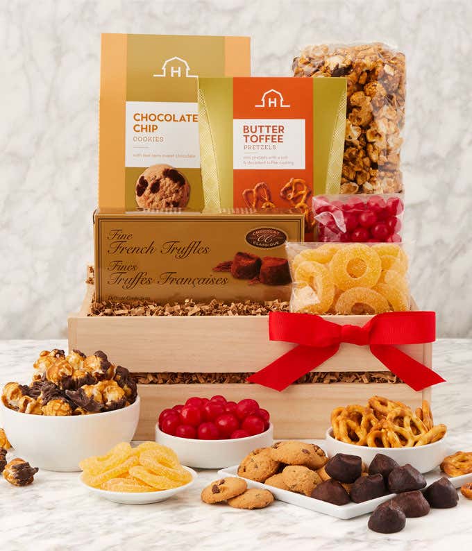 Wooden gift box with a red bow filled with sweet treats, including chocolate chip cookies, butter toffee pretzels, caramel popcorn, French truffles, cherry sours, and dried fruit slices. In front, bowls and piles display each treat for sharing.