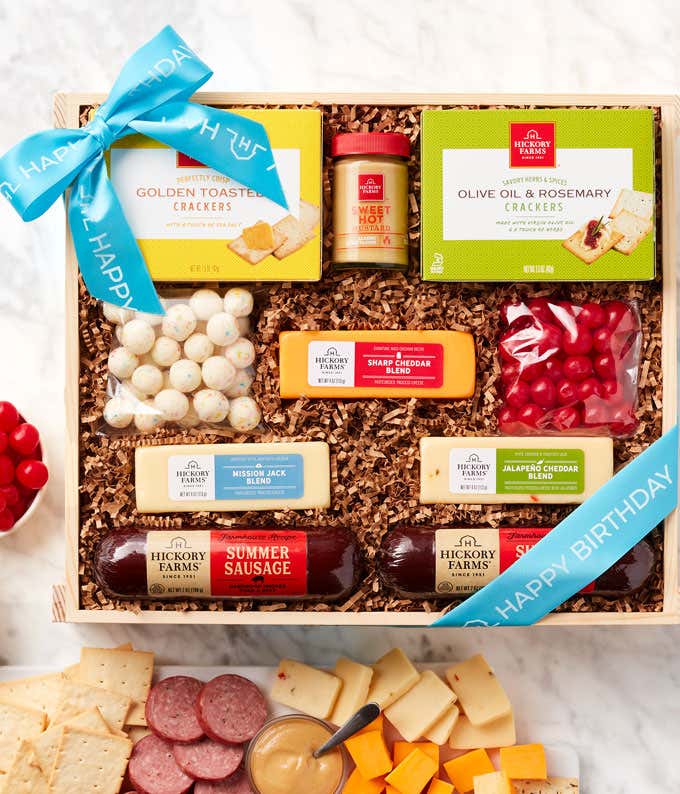 Wooden birthday gift box with a blue ribbon, filled with summer sausages, assorted cheeses, two types of crackers, mustard, white chocolate-covered treats, cherry sours, and a birthday cake bites. A serving board in front displays sausage and cheese.