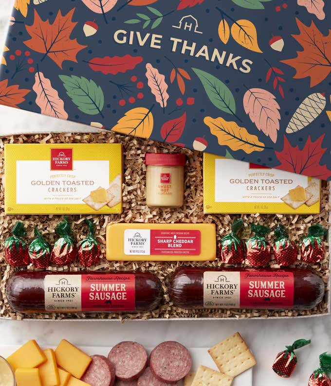 Gift box with a fall Give Thanks design, golden toasted crackers, three summer sausages, two cheese blocks, a small jar of sweet hot mustard, and assorted chocolate candies, with a serving tray displaying sliced sausage, cheese cubes, and crackers.