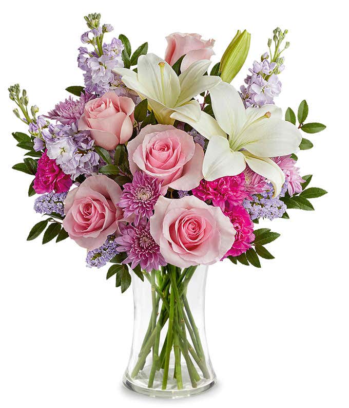 Image of a Mother's Day arrangement featuring Pink Roses, White Asiatic Lilies, Hot Pink Carnations, Lavender Stock, Lavender Cushion Spray Chrysanthemums, floral greenery, all beautifully presented in a metallic lavender vase.