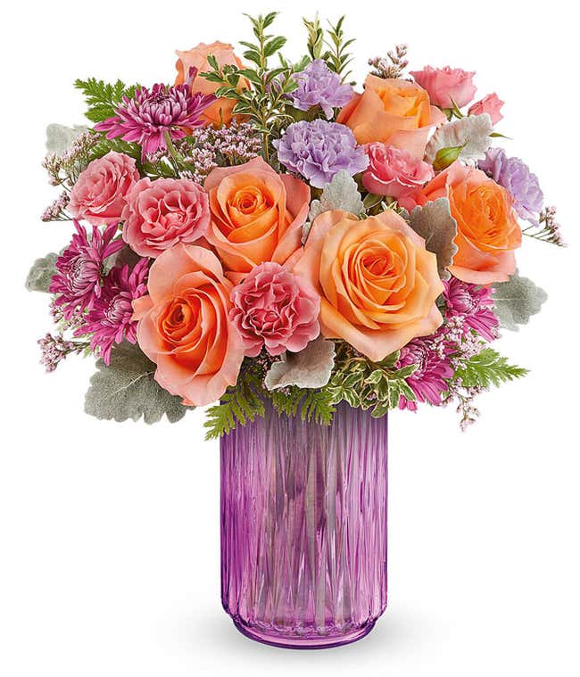 Vibrant floral arrangement featuring orange roses, pink miniature carnations, lavender carnations, pink cushion spray chrysanthemums, dusty miller, leatherleaf fern, and pink statice in a textured pink glass vase.