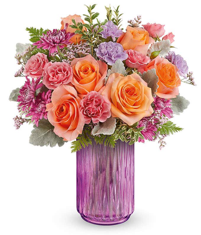 Vibrant floral arrangement featuring orange roses, pink miniature carnations, lavender carnations, pink cushion spray chrysanthemums, dusty miller, leatherleaf fern, and pink statice in a textured pink glass vase.