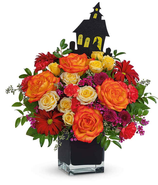 Halloween-themed flower arrangement with orange and yellow roses, red daisies, and pink filler flowers in a black cube vase, accented by greenery and topped with a black haunted house decoration.