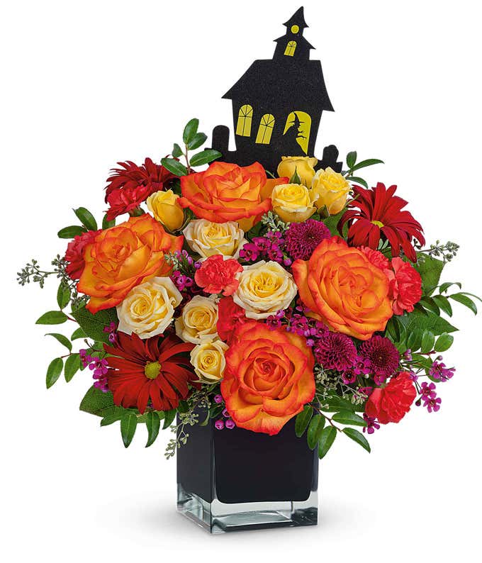 Halloween-themed flower arrangement with orange and yellow roses, red daisies, and pink filler flowers in a black cube vase, accented by greenery and topped with a black haunted house decoration.