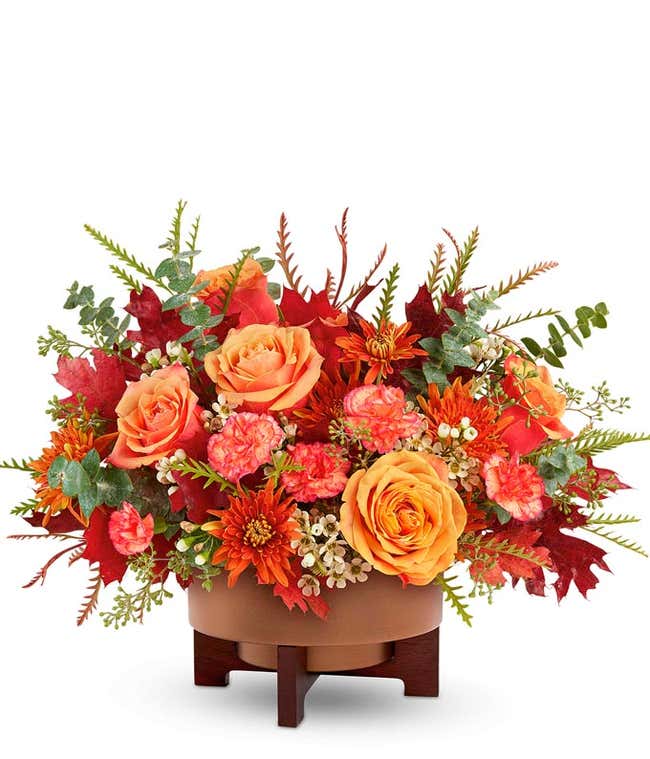 Fall flower arrangement with peach roses, orange carnations, rust-colored daisy chrysanthemums, white waxflower, and assorted autumn leaves in a brown ceramic container on a wooden stand.