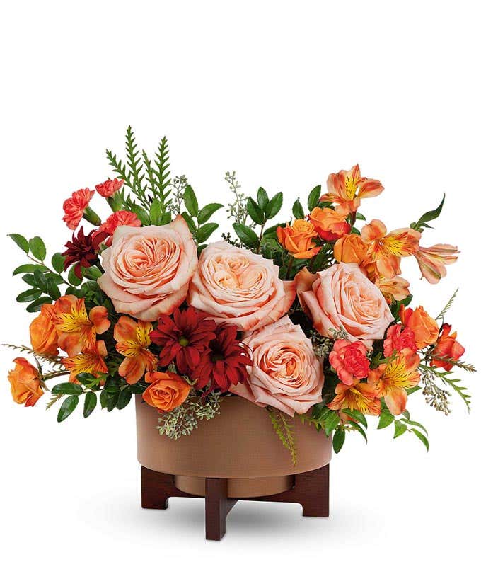 Fall flower arrangement with light orange roses, orange alstroemeria, miniature red carnations, and rust-colored daisy spray chrysanthemums, displayed in a brown ceramic container with a wooden stand.