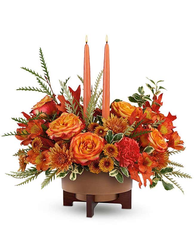 Fall centerpiece with orange roses, orange spray roses, red carnations, bronze cushion spray chrysanthemums, fall leaves, and greenery, arranged around two orange taper candles in a brown ceramic planter with wooden stand.