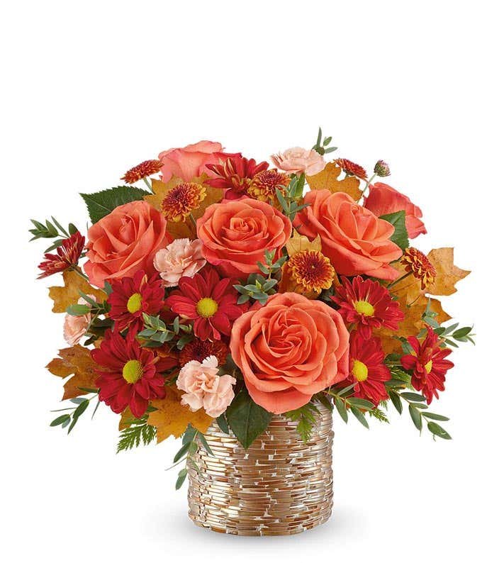 Fall floral arrangement with orange roses, red daisy poms with yellow centers, rust chrysanthemums, light peach carnations, and autumn leaves in a textured gold-glass vase.
