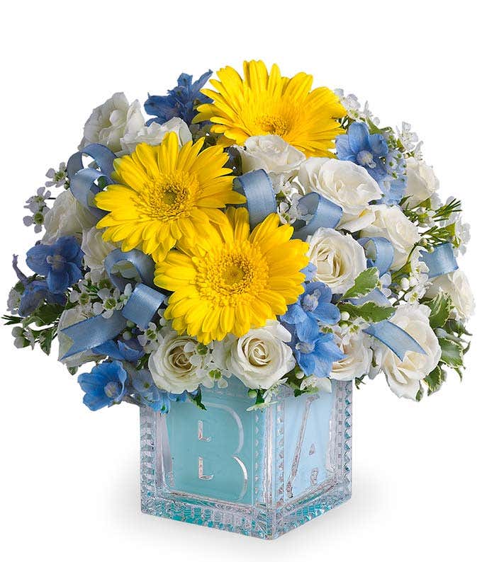 A charming arrangement featuring yellow daisies, white spray roses, and blue delphinium, elegantly presented in a baby blue block vase with a decorative blue ribbon. This mix offers a cheerful and serene display.