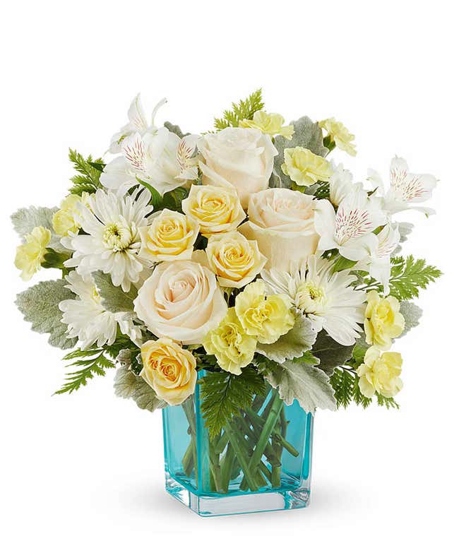 Cheerful floral arrangement with creamy white roses, soft yellow spray roses, white alstroemeria, white mums, and yellow carnations in a vibrant blue glass vase, perfect gift for loved ones.