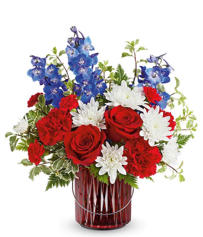Red, white, and blue floral arrangement with red roses, white chrysanthemums, red carnations, blue delphinium, and assorted greenery in a red glass vase.