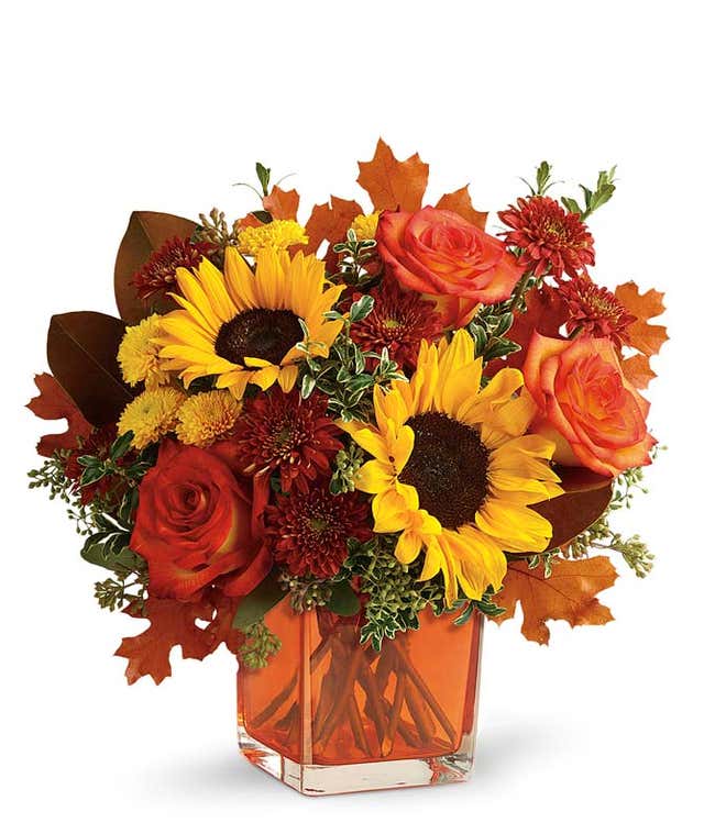 A vibrant floral arrangement featuring orange roses, yellow sunflowers, gold cushion poms, and rust cushion mums, accented with seeded eucalyptus, oregonia, and magnolia leaves, all beautifully presented in a keepsake orange cube vase.