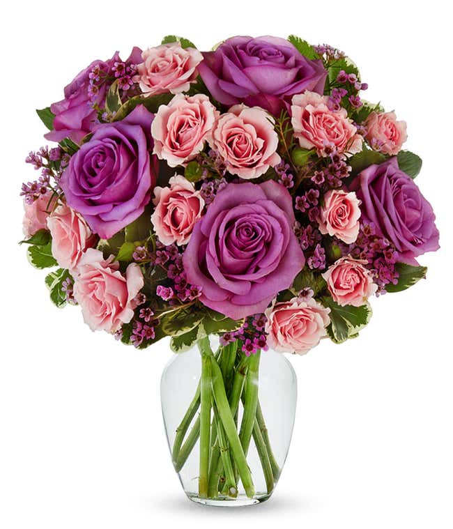 A vibrant bouquet of lavender roses, pink spray roses, and purple waxflower accented with lush greenery, arranged in a clear glass vase.