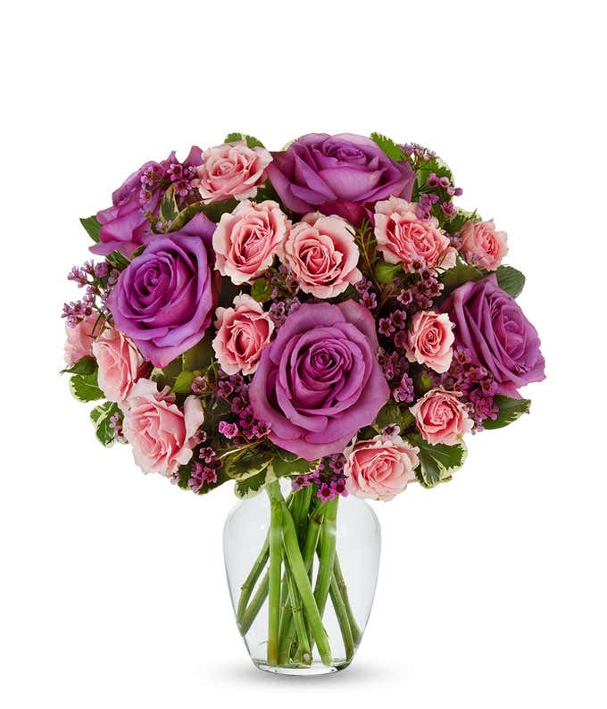 A bouquet of lavender roses, pink spray roses, and purple filler flowers arranged in a clear glass vase with greenery.