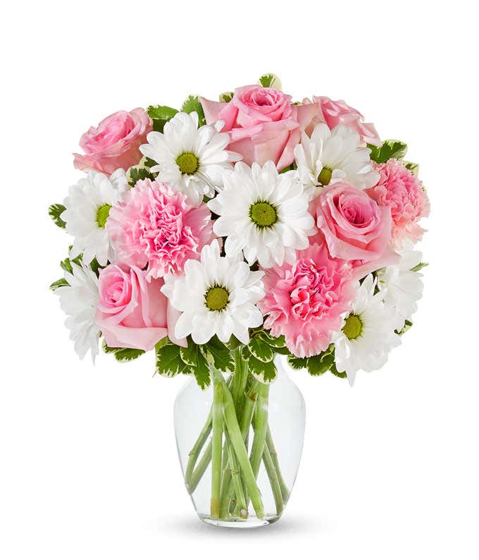 A bouquet of pink roses, pink carnations, and white daisy poms with greenery in a clear glass vase.