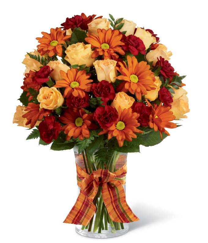 Clear glass vase filled with orange daisy poms, peach roses, and red mini carnations, accented with greenery and tied with an orange plaid ribbon.