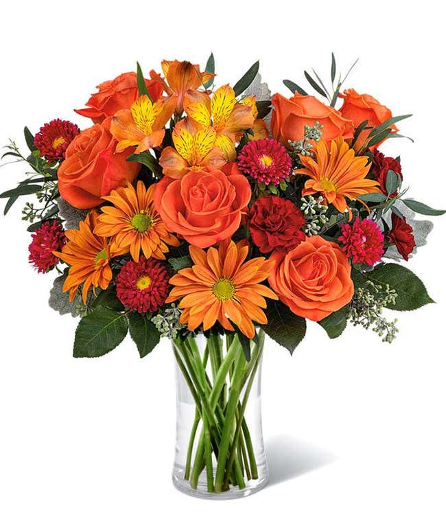 A vibrant floral arrangement featuring orange roses, orange alstroemeria, bronze daisies, red Matsumoto asters, maroon mini carnations, and seasonal greenery, complemented by a personal card message, all beautifully presented in a clear cylinder vase.