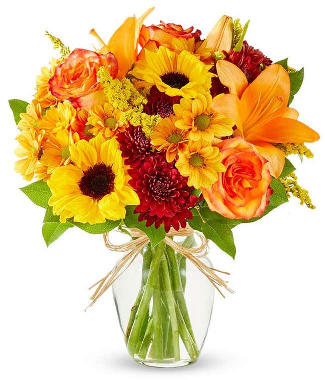 Bright autumn bouquet with sunflowers, orange lilies, orange roses, burgundy poms, yellow solidago, and butterscotch daisies in a clear glass vase tied with a raffia bow.