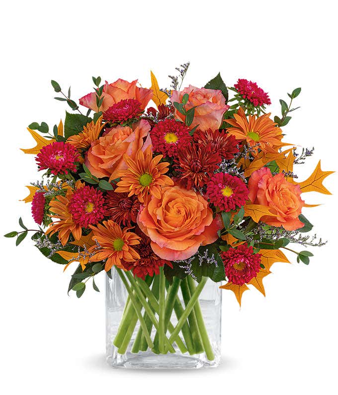 Fall flower arrangement with orange roses, red Matsumoto asters, bronze daisy poms, red mums, greenery, purple filler flowers, and autumn oak leaves in a clear glass cube vase.
