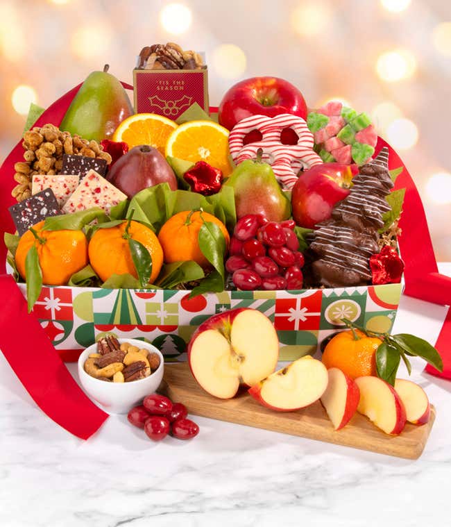 Festive holiday gift basket filled with assorted fruits, including apples, pears, and oranges. The basket also contains chocolate-covered pretzels, peppermint bark, caramel popcorn, mixed nuts, and holiday-themed candies, decorated with a red ribbon.