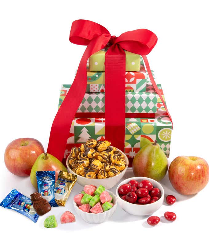 A festive stack of gift boxes wrapped with a red bow, filled with apples, pears, caramel popcorn, chocolate cherries, and red and green gummy candies. 