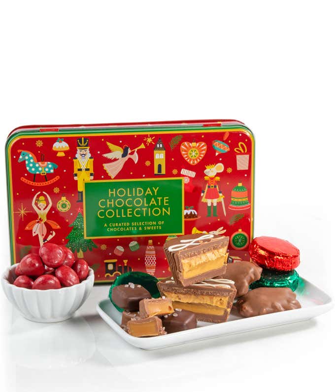 Holiday chocolate collection gift tin with festive illustrations, shown with assorted chocolates, chocolate-covered caramels, peanut butter cups, and a small bowl of red candy-coated chocolates on a white tray.