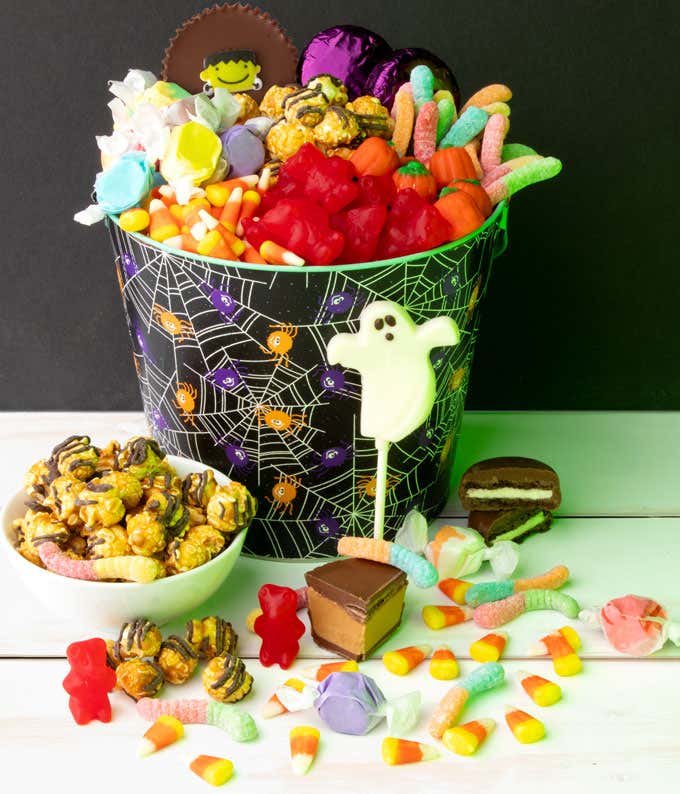 Halloween candy bucket filled with popcorn, candy corn, gummy worms, gummy bears, taffy, chocolate treats, and topped with a ghost lollipop, with additional candies and popcorn scattered around the base.