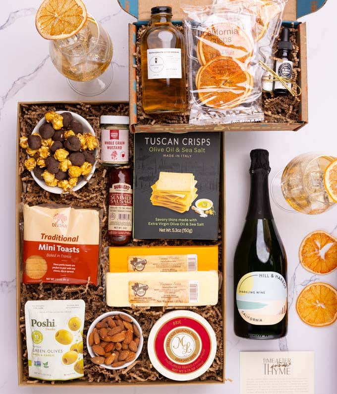 A bottle of Sparkling Wine next to a box holding three cheeses, sausage, crackers, olives, almonds, popcorn & mustard.  The cocktail box contains thyme-cucumber syrup, celery bitters, grapefruit oil mist, dehydrated grapefruit slices, & recipe card.